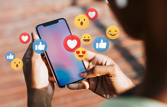 social-media-image Hands holding a smartphone with social media emojis floating around the screen